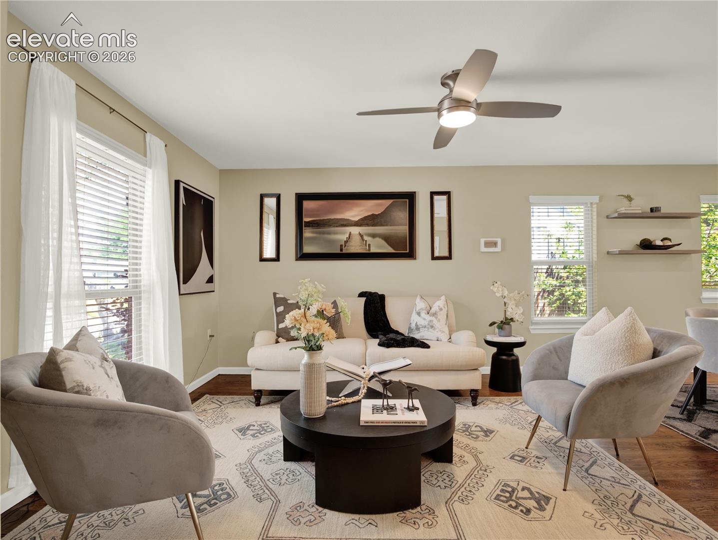 Living area featuring wood-finish flooring, a contemporary brushed-metal ceiling fan with integrated lighting, dual wall-mounted floating shelves, and multiple windows with horizontal blinds