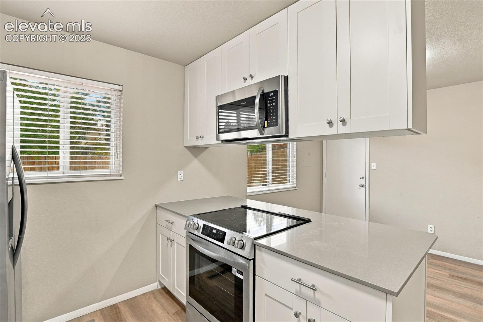 Kitchen featuring white shaker-style cabinetry, stainless steel appliances, neutral stone-finish countertops, and wood-finish flooring
