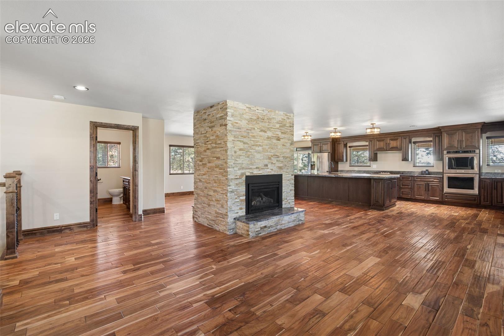 Stacked stone fireplace with a black insert and stone hearth, wood-finish flooring, and kitchen featuring custom cabinetry, integrated double wall ovens, and a large island