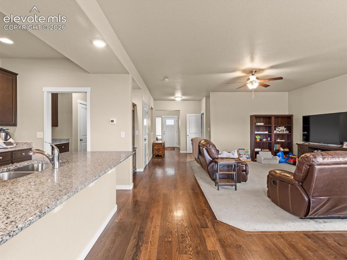 Open-concept living space featuring wood-finish flooring extending into the kitchen, recessed ceiling lighting, and a kitchen island with a double basin sink and light-toned countertop