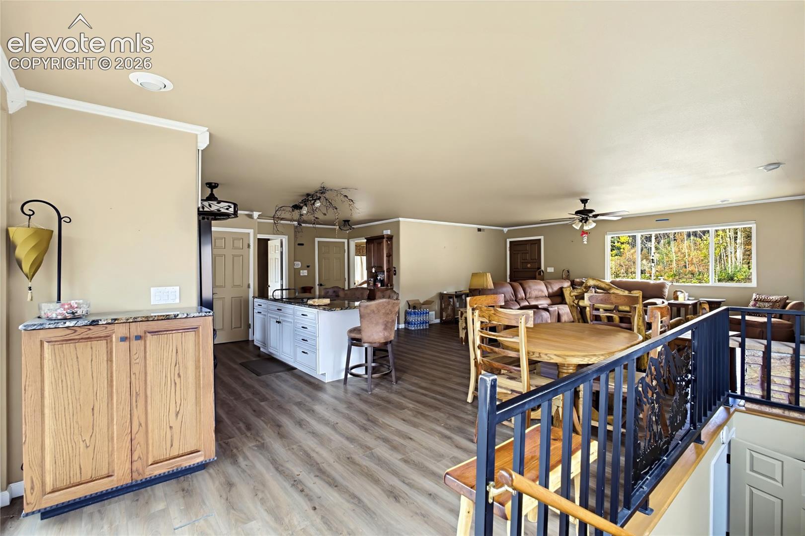 Open-concept interior featuring wood-finish flooring, a kitchen island with stone countertop, recessed lighting, ceiling fans, and black metal stair railing
