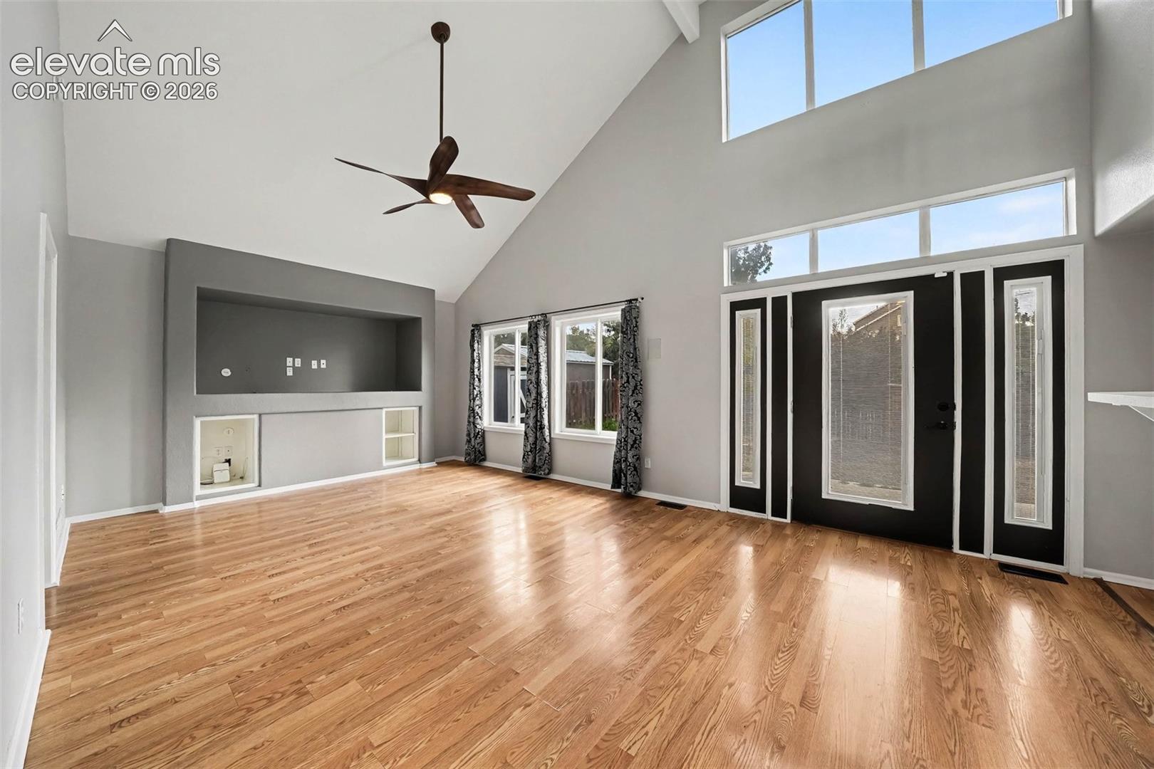 Vaulted ceiling with an integrated ceiling fan, wood-finish flooring, built-in entertainment niche, and a panel-glass entry door with sidelights and transom windows