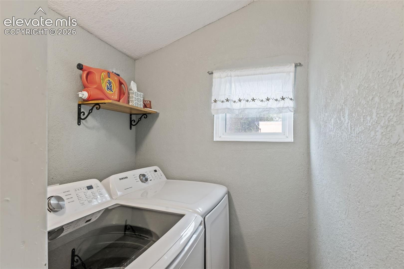 Dedicated laundry area featuring a window, textured walls, and a built-in wooden shelf with decorative metal brackets