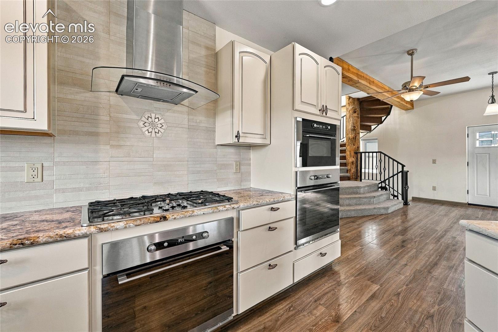 Kitchen featuring off-white cabinetry, stone-finish countertops, stainless steel appliances including a wall-mounted glass range hood, horizontal tiled backsplash, and wood-finish flooring
