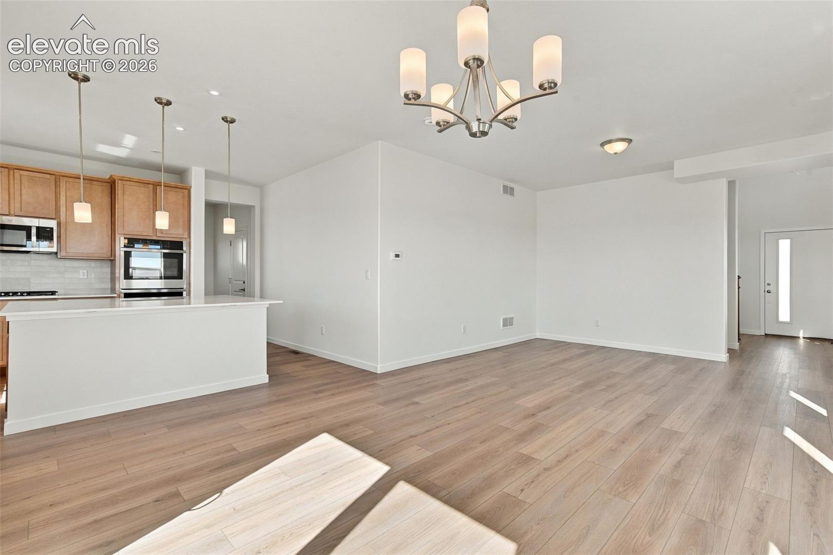 Open-concept living space featuring wood-finish flooring, a kitchen island with pendant lighting, stainless steel kitchen appliances, and an entry door with a frosted glass insert
