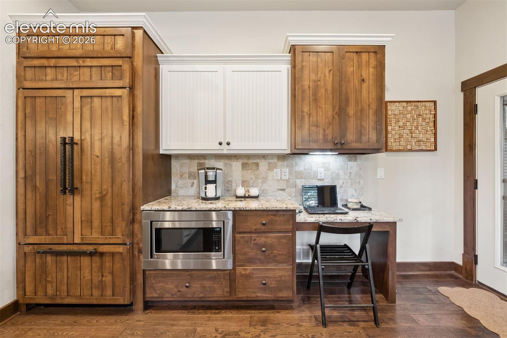 Custom wood-paneled refrigerator, natural stone backsplash, light-toned stone countertop, built-in microwave, and wood-finish flooring