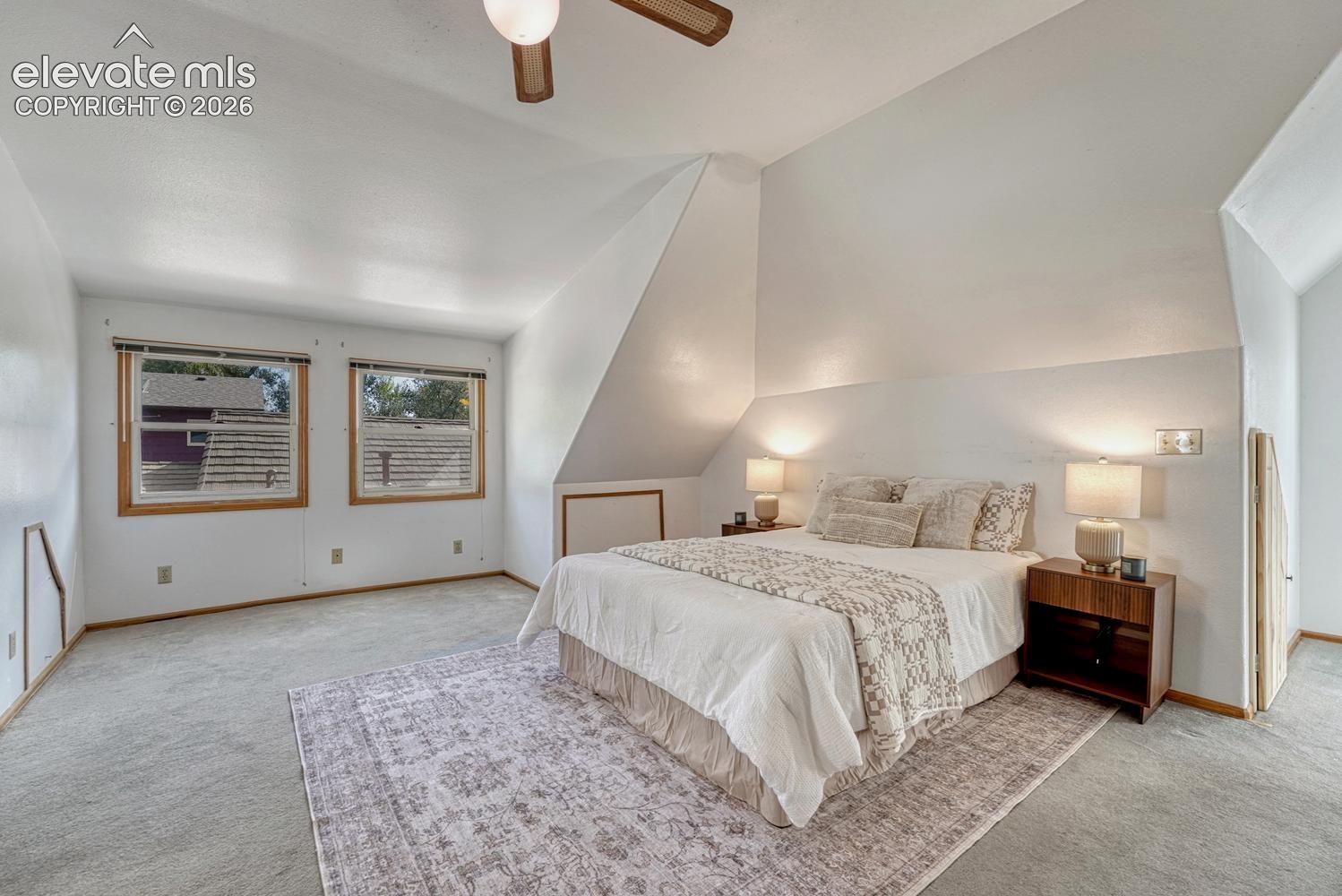 Sloped ceiling bedroom featuring light-toned carpeting, two windows with wood-finish trim, and a ceiling fan