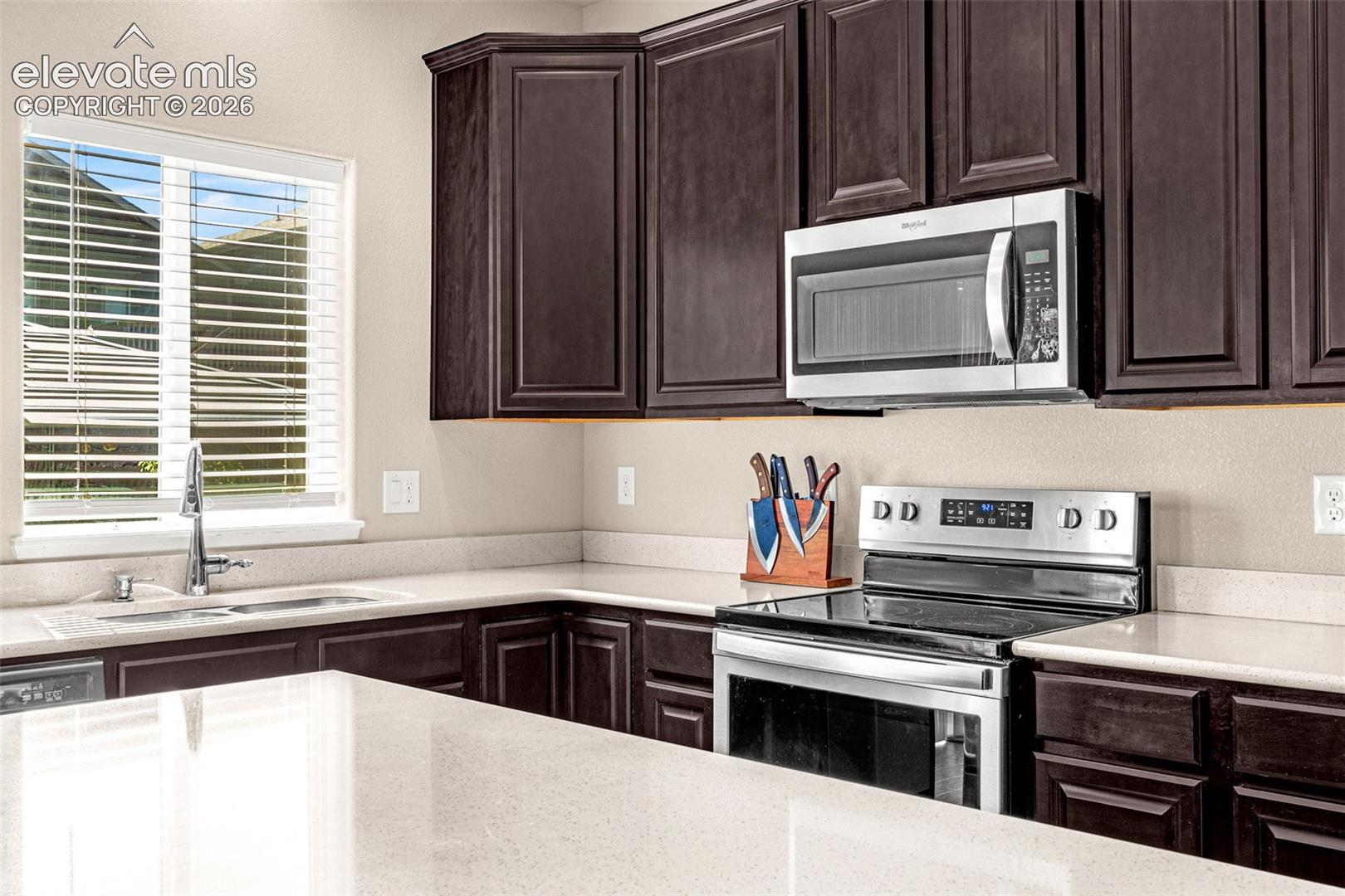 Dark wood-finish cabinetry, light-toned countertops, stainless steel appliances, and an undermount sink with a gooseneck faucet