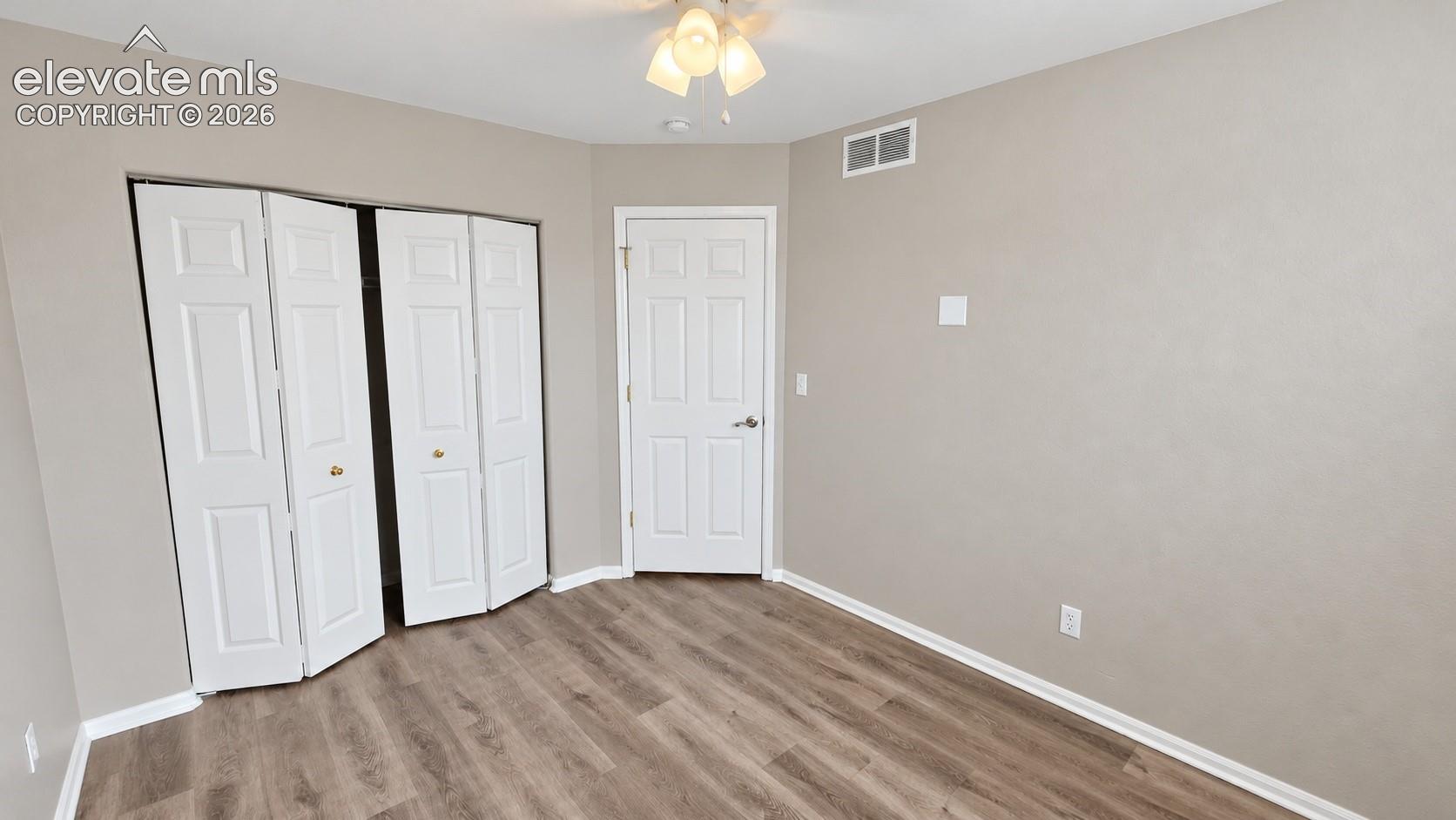 Neutral-toned interior featuring wood-finish flooring, a bi-fold closet, a solid-panel door, and a three-bulb flush-mount light fixture
