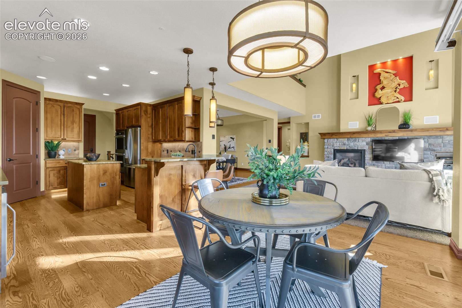 Open-concept area featuring wood-finish flooring, custom cabinetry with a center island, stone-surround fireplace with a timber mantel, and decorative wall niches
