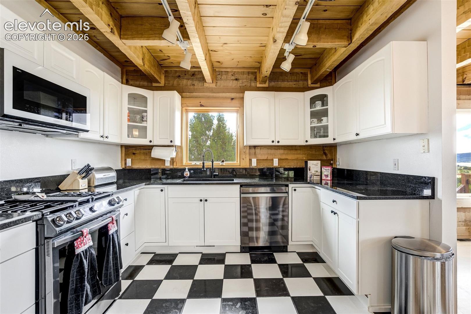 U-shaped kitchen featuring white cabinetry, black countertops, stainless steel appliances, a black-and-white checkered floor, and wood plank ceiling with exposed beams