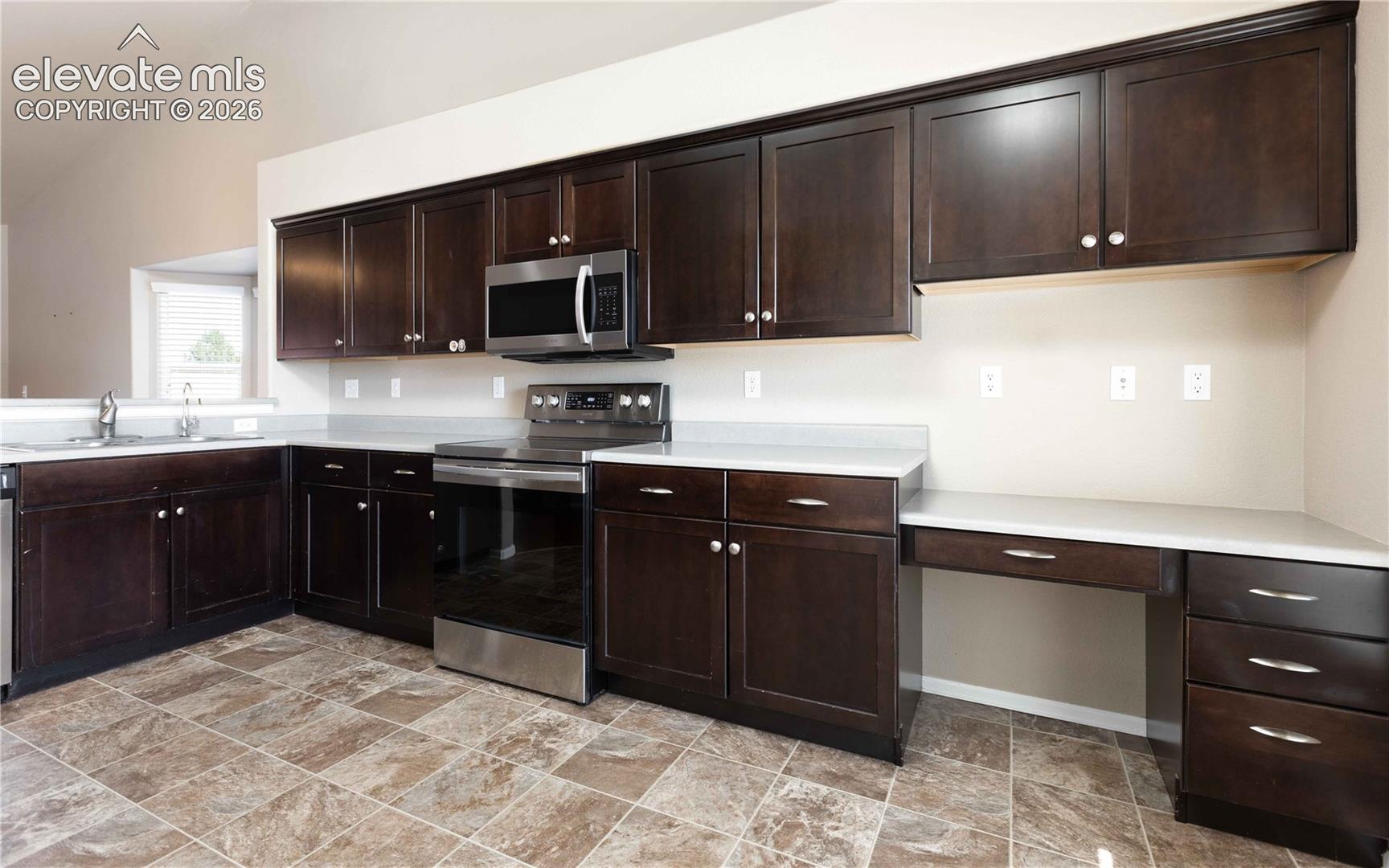 Kitchen featuring dark wood cabinetry, stainless steel appliances, neutral stone-finish countertops, tile flooring, and a built-in desk area