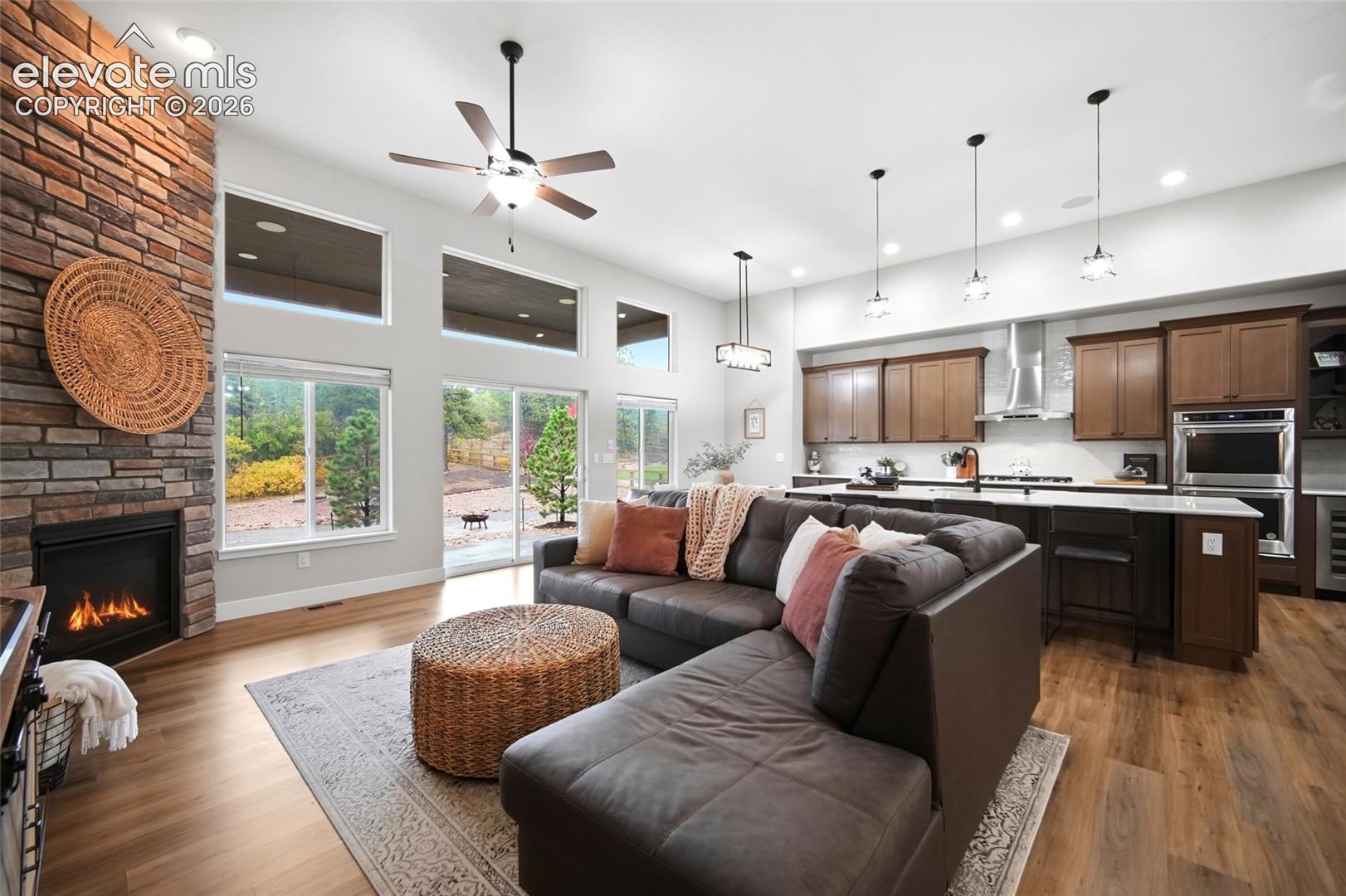 Open-concept interior featuring wood-finish flooring, a floor-to-ceiling stone fireplace, high ceilings with clerestory windows, and a kitchen equipped with dark wood cabinetry and a central island with integrated seating