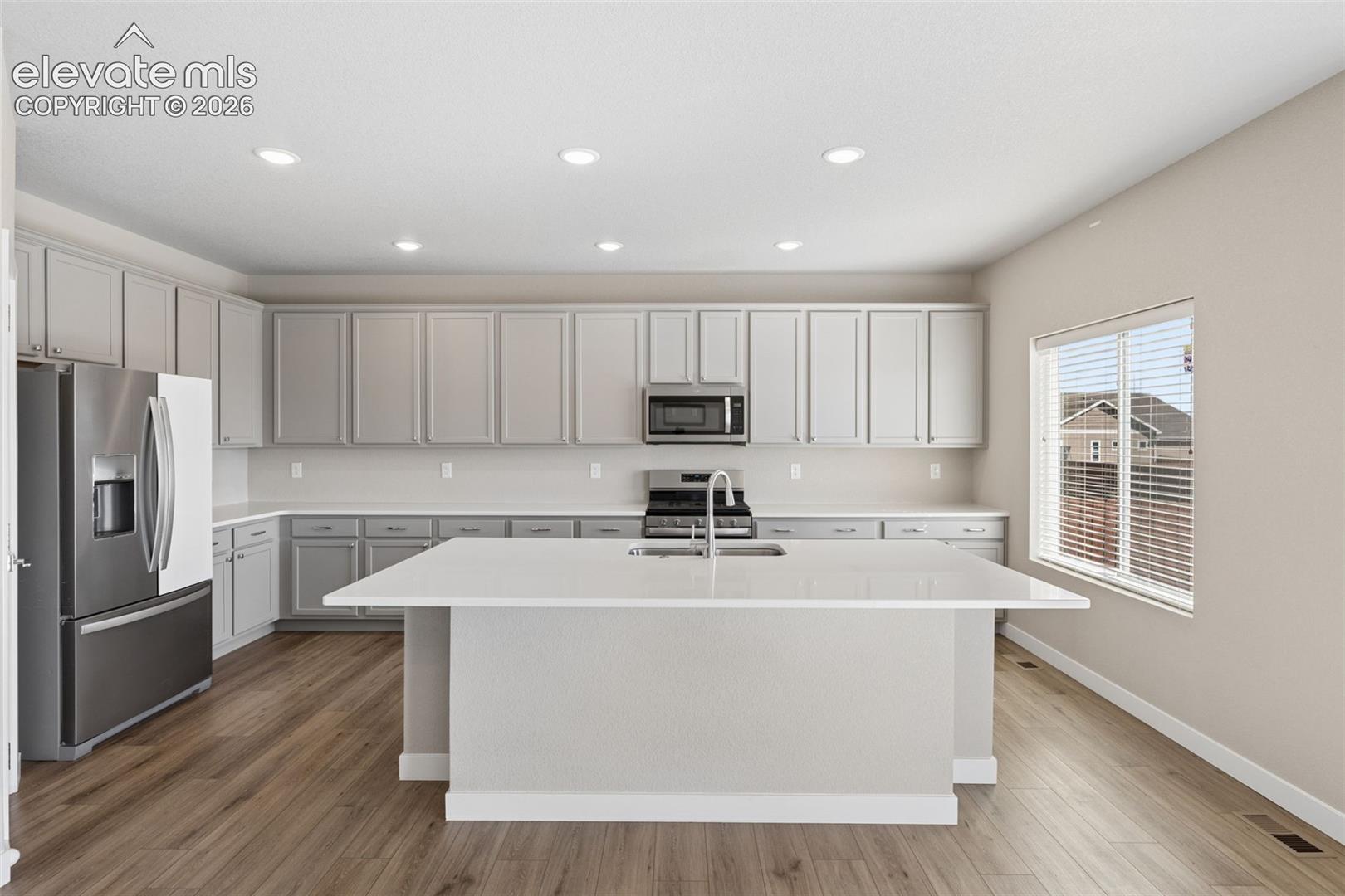 Kitchen featuring light-toned cabinetry, quartz countertops, a central island with an integrated sink, stainless steel appliances, and wood-finish flooring