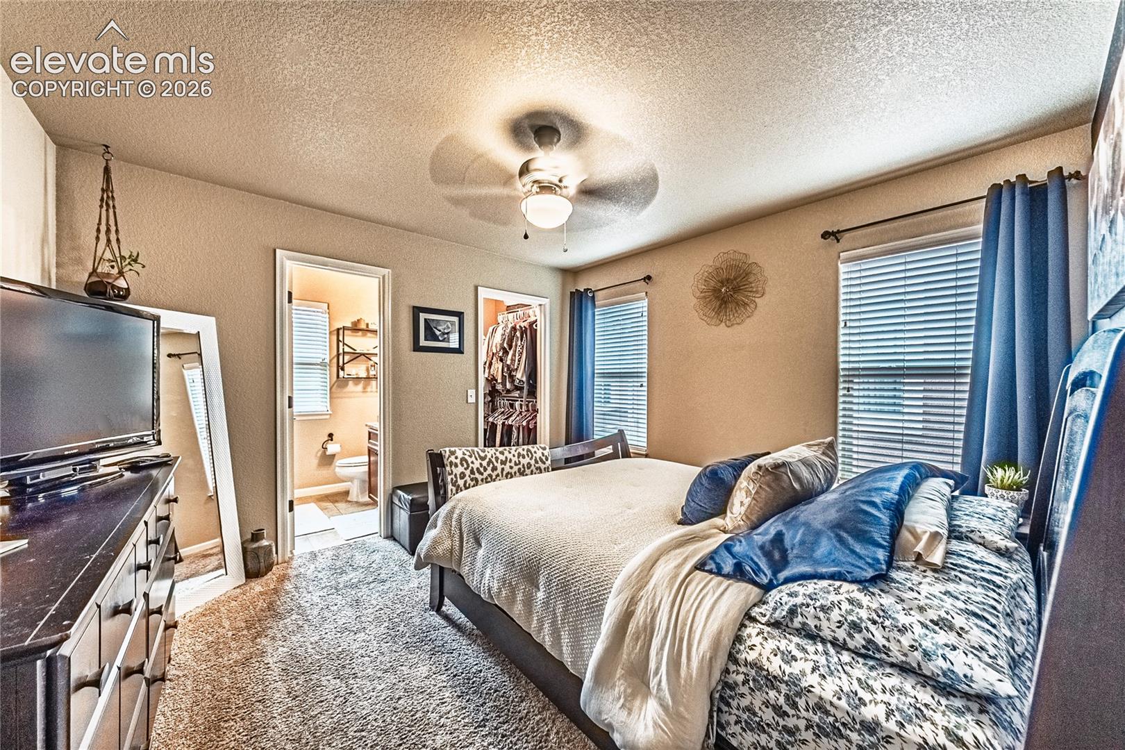 Carpeted bedroom featuring a textured ceiling, a walk in closet, a ceiling fan, a textured wall, and ensuite bathroom