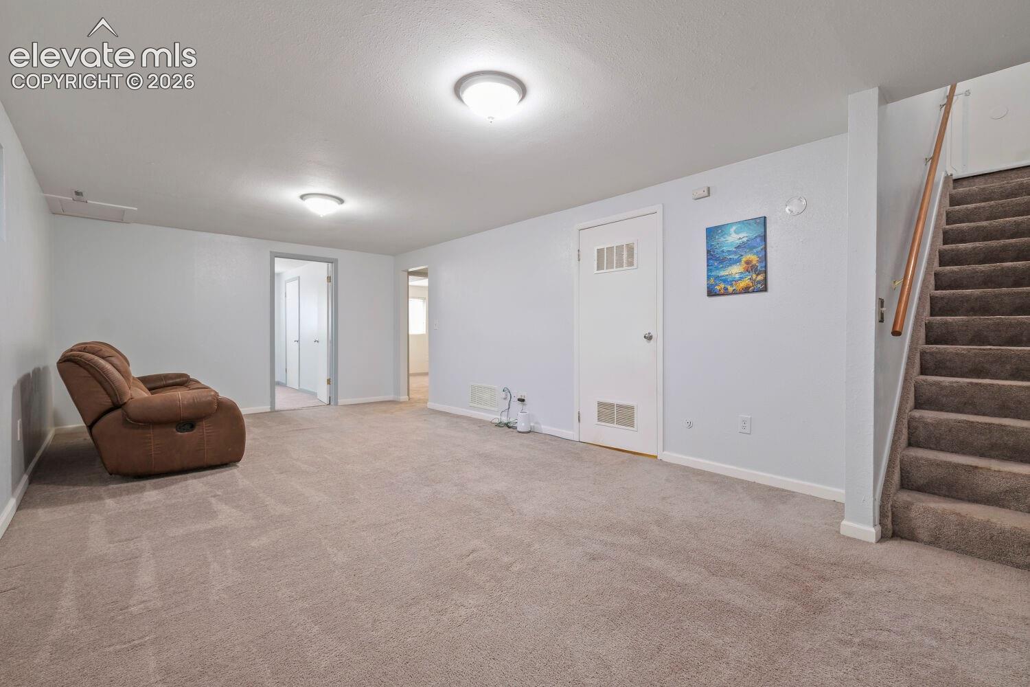 Spacious carpeted room featuring white walls, flush-mount ceiling lighting, a white door with louvered panels, and a carpeted staircase with a wood handrail