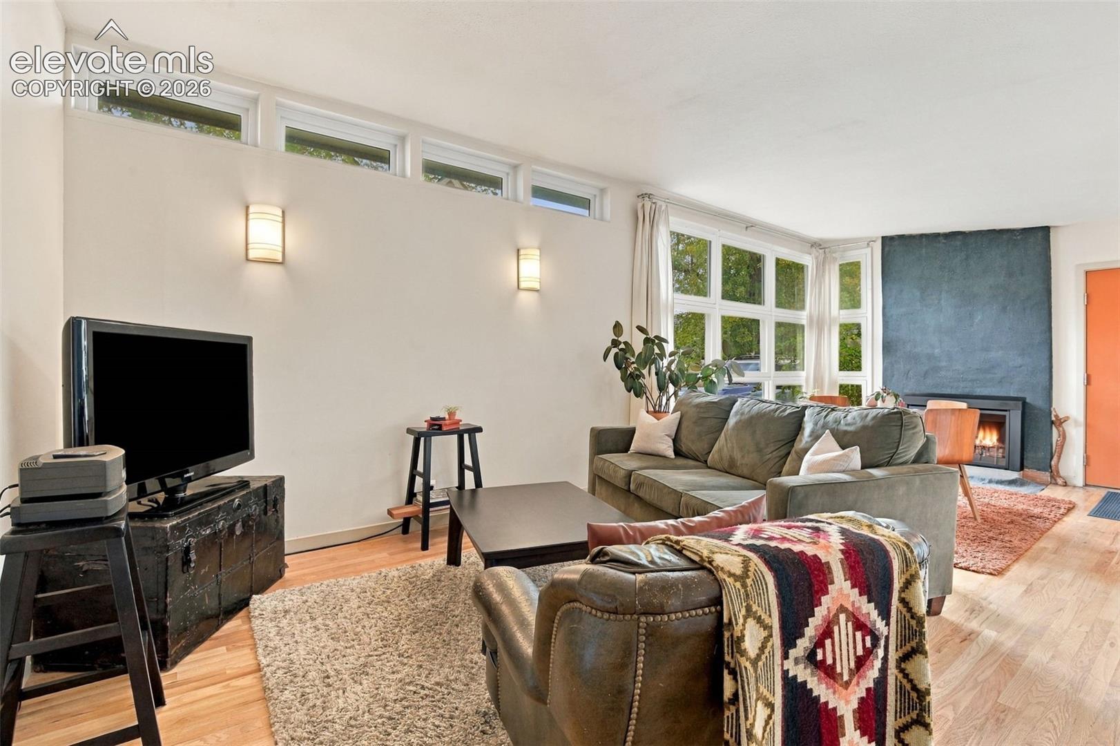 Living space featuring clerestory windows, wood-finish flooring, a floor-to-ceiling dark stone-finish fireplace surround, and wall-mounted light fixtures