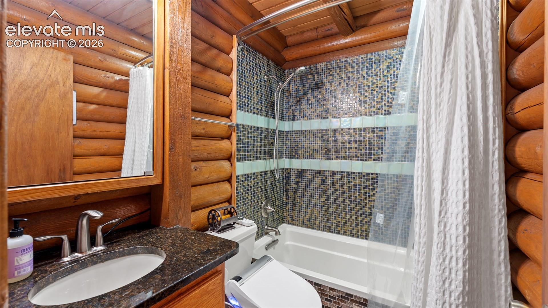 Full-round log walls, custom glass tile shower surround with accent stripe, dark stone-finish countertop, undermount oval sink, and polished chrome faucet fixtures