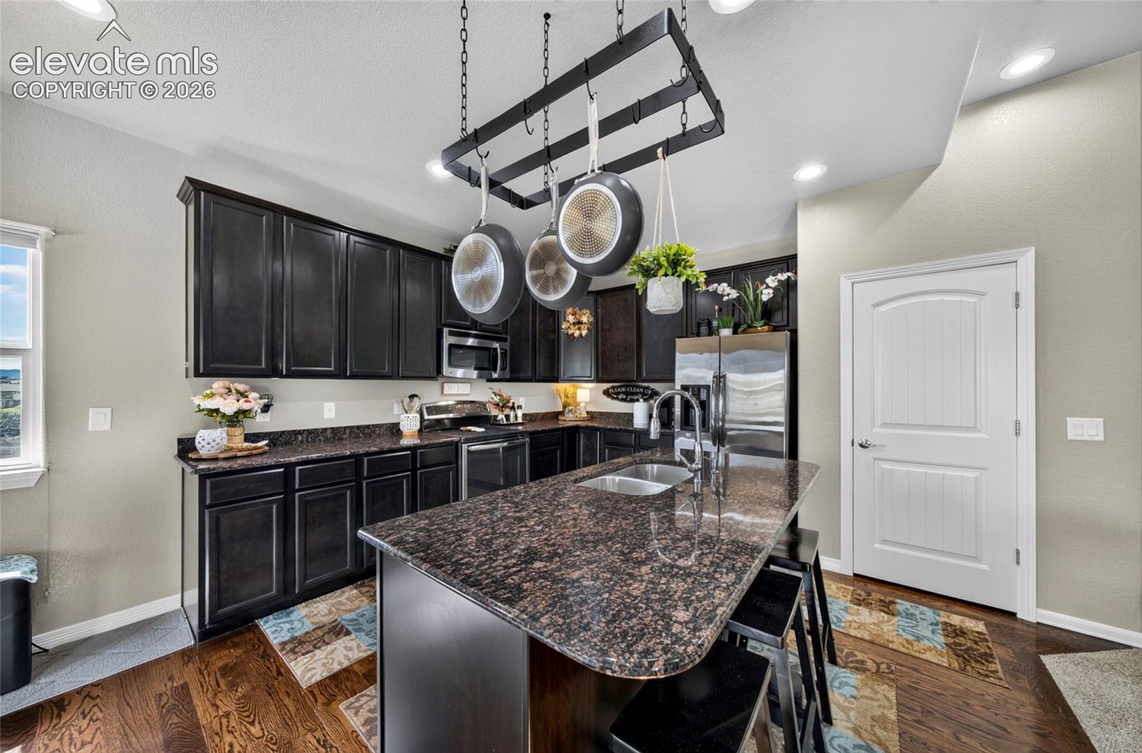 Kitchen featuring dark cabinetry, expansive stone countertops, a central island with an integrated stainless steel dual-basin sink, and a suspended metal pot rack