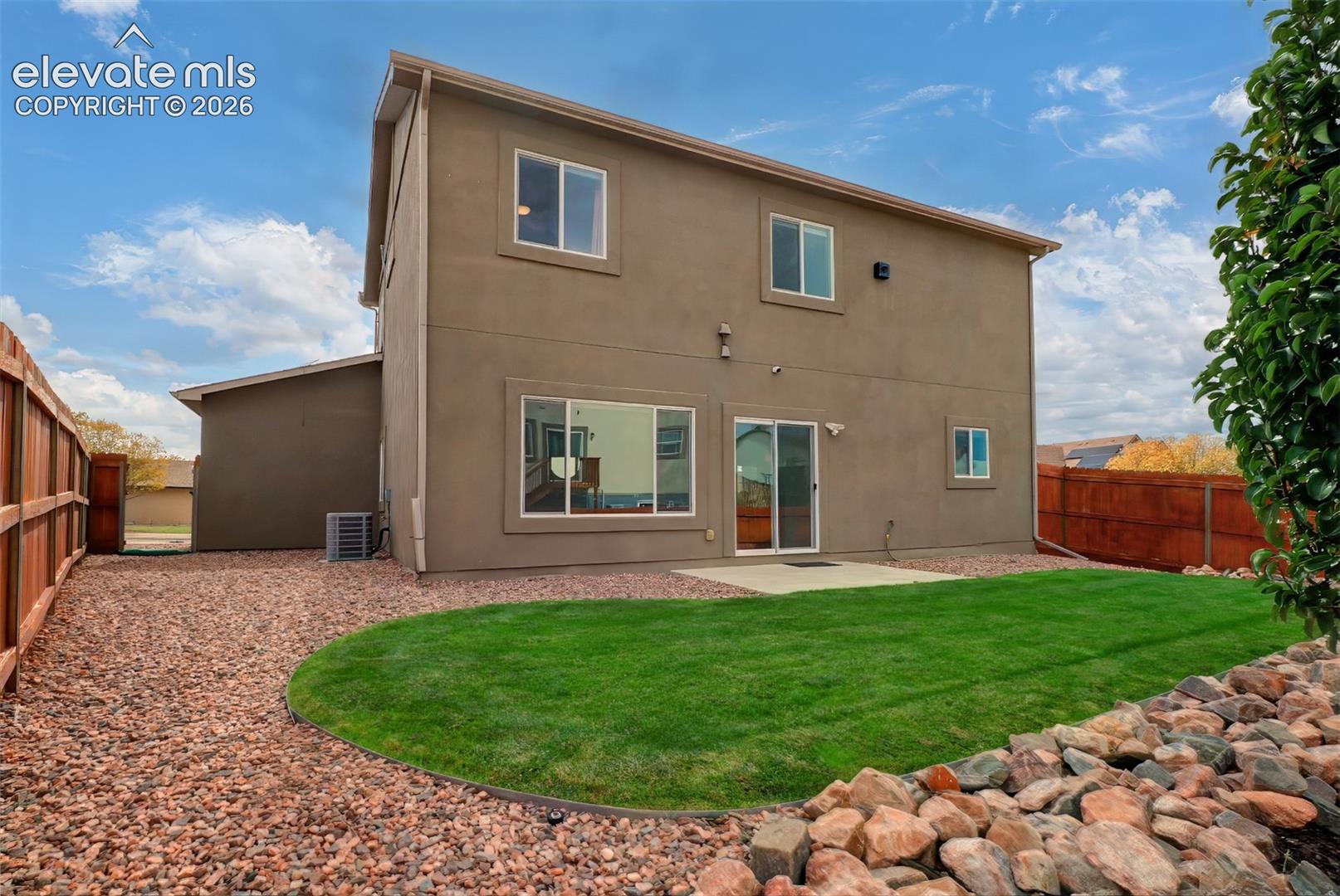 Two-story stucco exterior featuring a concrete patio, gravel landscaping, a manicured lawn, and wooden privacy fencing