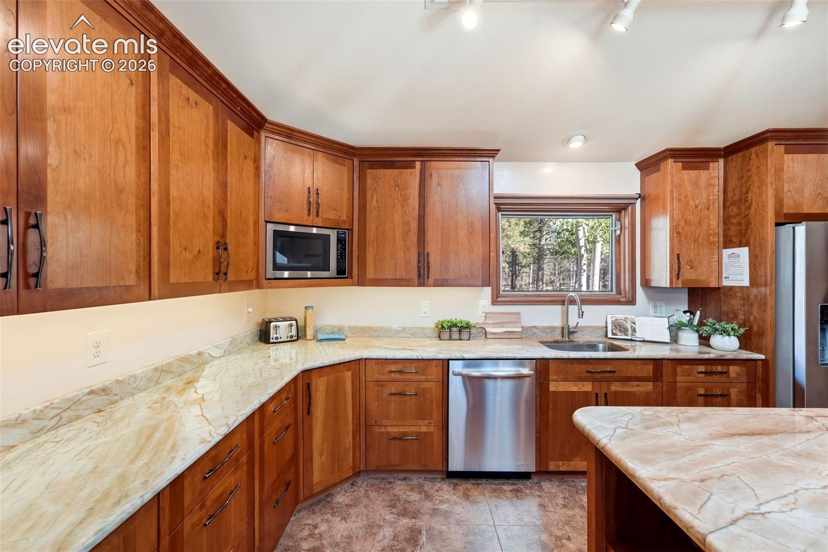 Wood cabinetry with crown molding, light-toned stone countertops, stainless steel appliances, a stainless steel sink with a gooseneck faucet, and track lighting