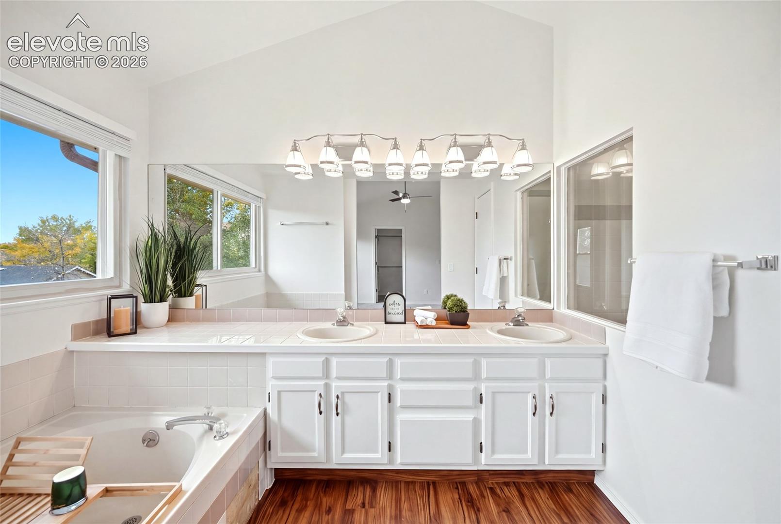 Vaulted ceiling, dual-sink vanity with tile countertop, wood-finish flooring, full-width wall mirror, and soaking tub with tile surround