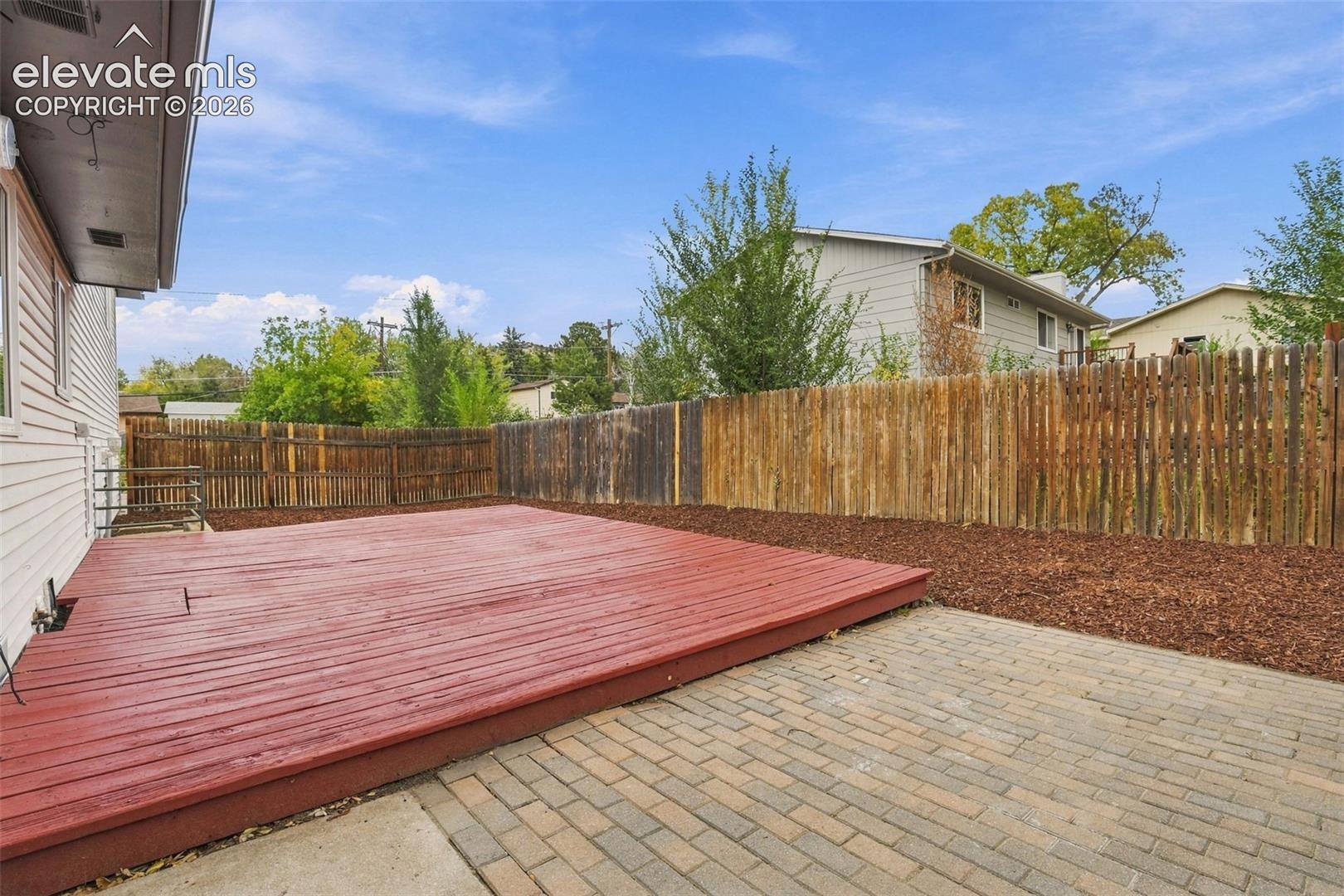 Outdoor living area featuring a wood deck, stone-paver patio, wood-slat perimeter fencing, and mulched landscaping