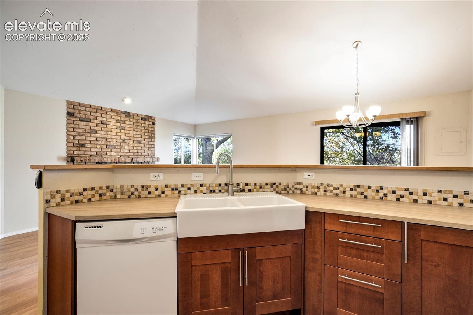 Kitchen featuring wood-finish cabinetry, farmhouse-style apron sink, mosaic tile backsplash, and brick fireplace accent wall