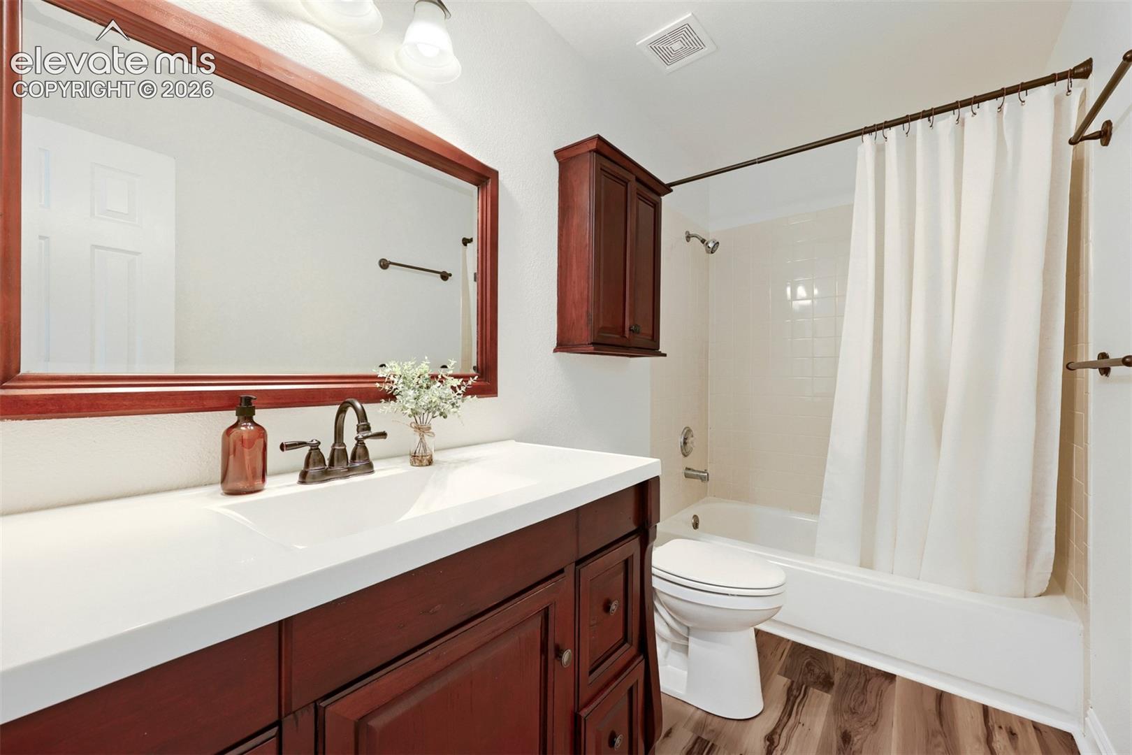 Wood-finish flooring, single vanity with a white countertop, large framed mirror, and a built-in shower/tub combination
