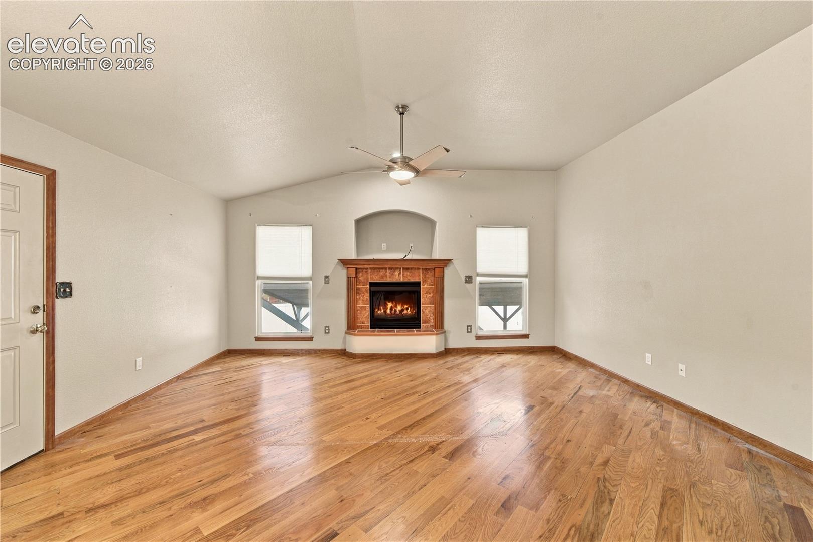 Living area featuring vaulted ceilings, wood-finish flooring, a central fireplace with a tiled surround, a recessed arched wall niche, and a ceiling fan