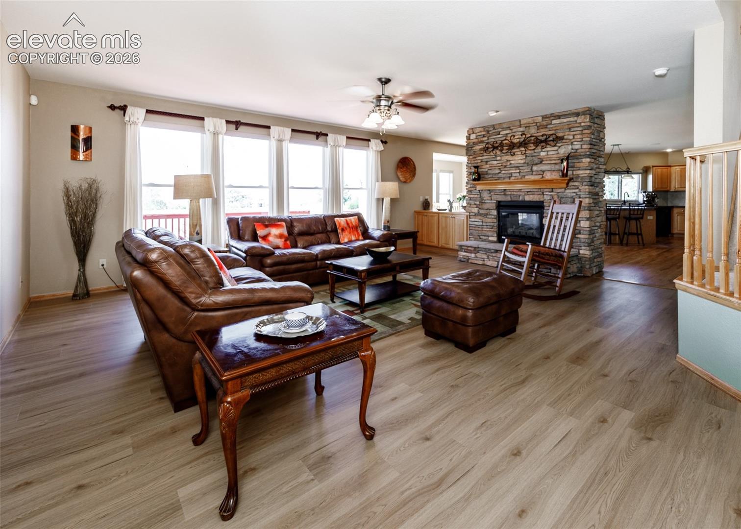 Wood-finish flooring, stone-clad fireplace with wood mantel, ceiling fan with integrated lighting, multiple large windows, and a wood railing