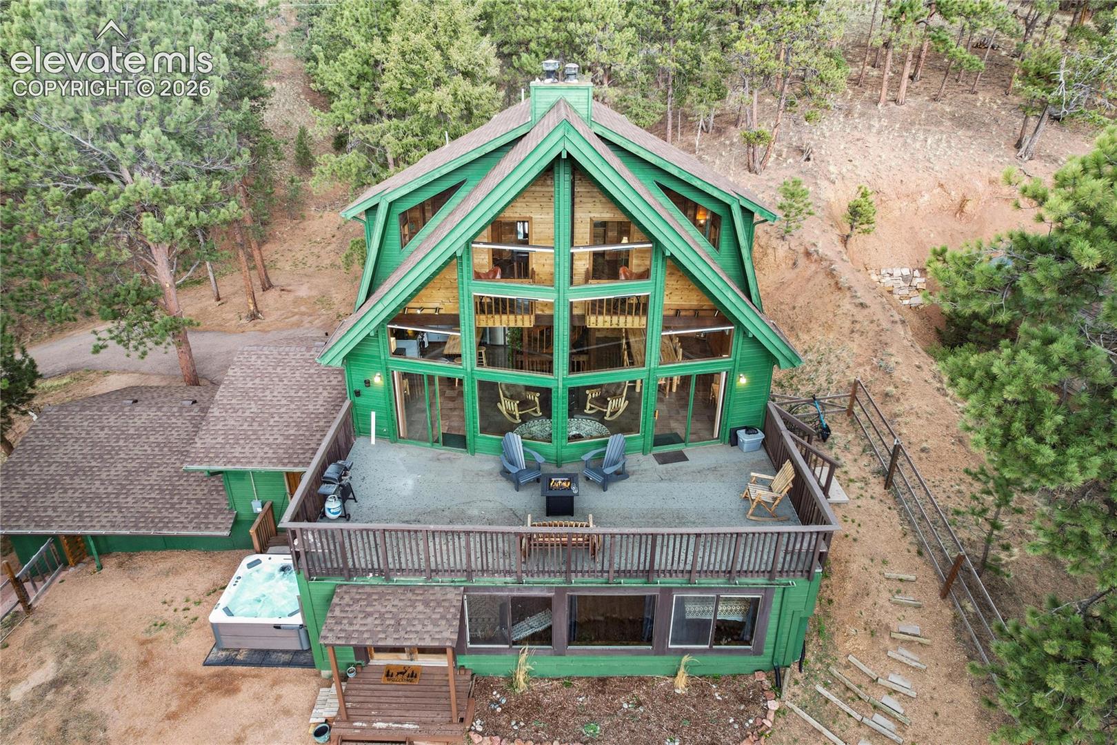 Multi-level A-frame residence featuring expansive floor-to-ceiling windows, wrap-around elevated deck, integrated hot tub, and wood-paneled siding situated within a wooded landscape