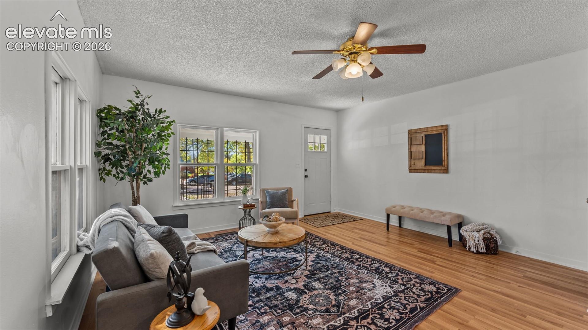 Bright and open living space featuring hardwood flooring, a ceiling fan with integrated lighting, and large windows offering natural light throughout