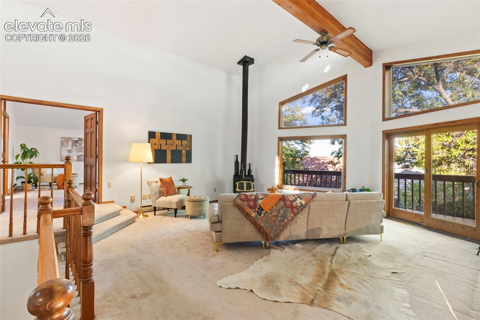 Vaulted ceilings featuring an exposed wood beam, a freestanding wood-burning stove with a chimney pipe, multi-level oversized windows, and wood-framed sliding glass doors