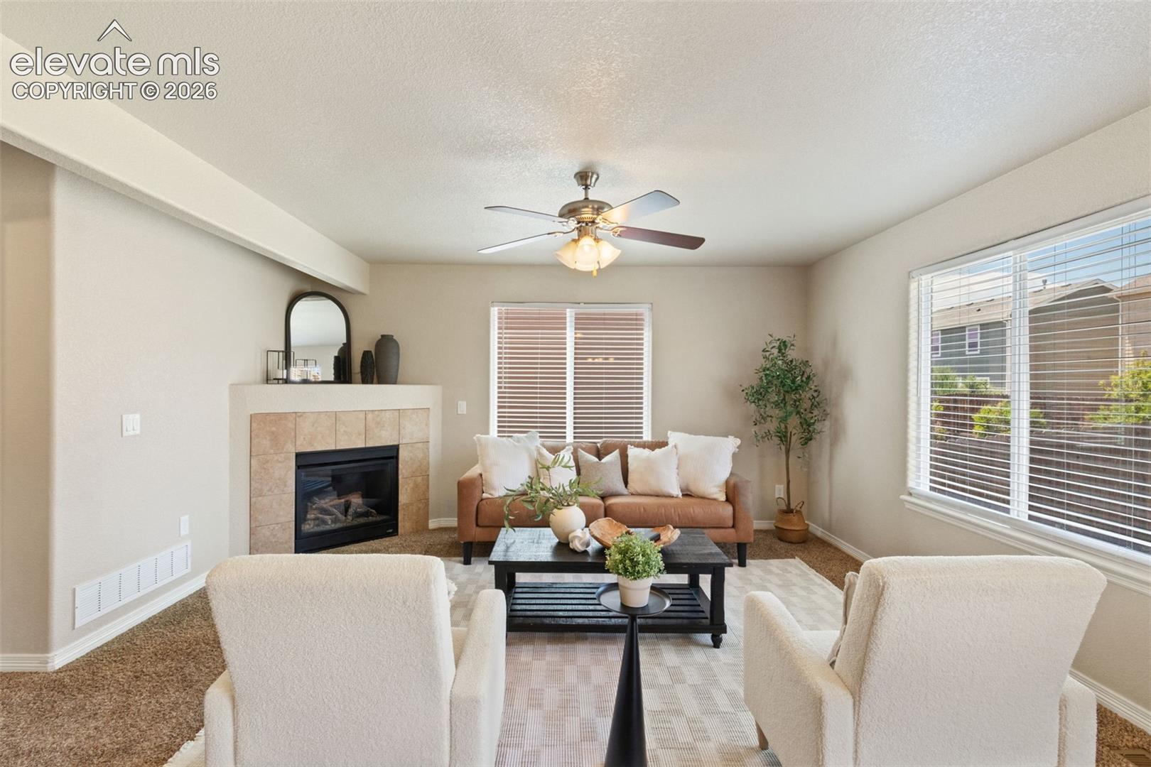 Living area with a tiled fireplace, large windows with blinds, and a ceiling fan