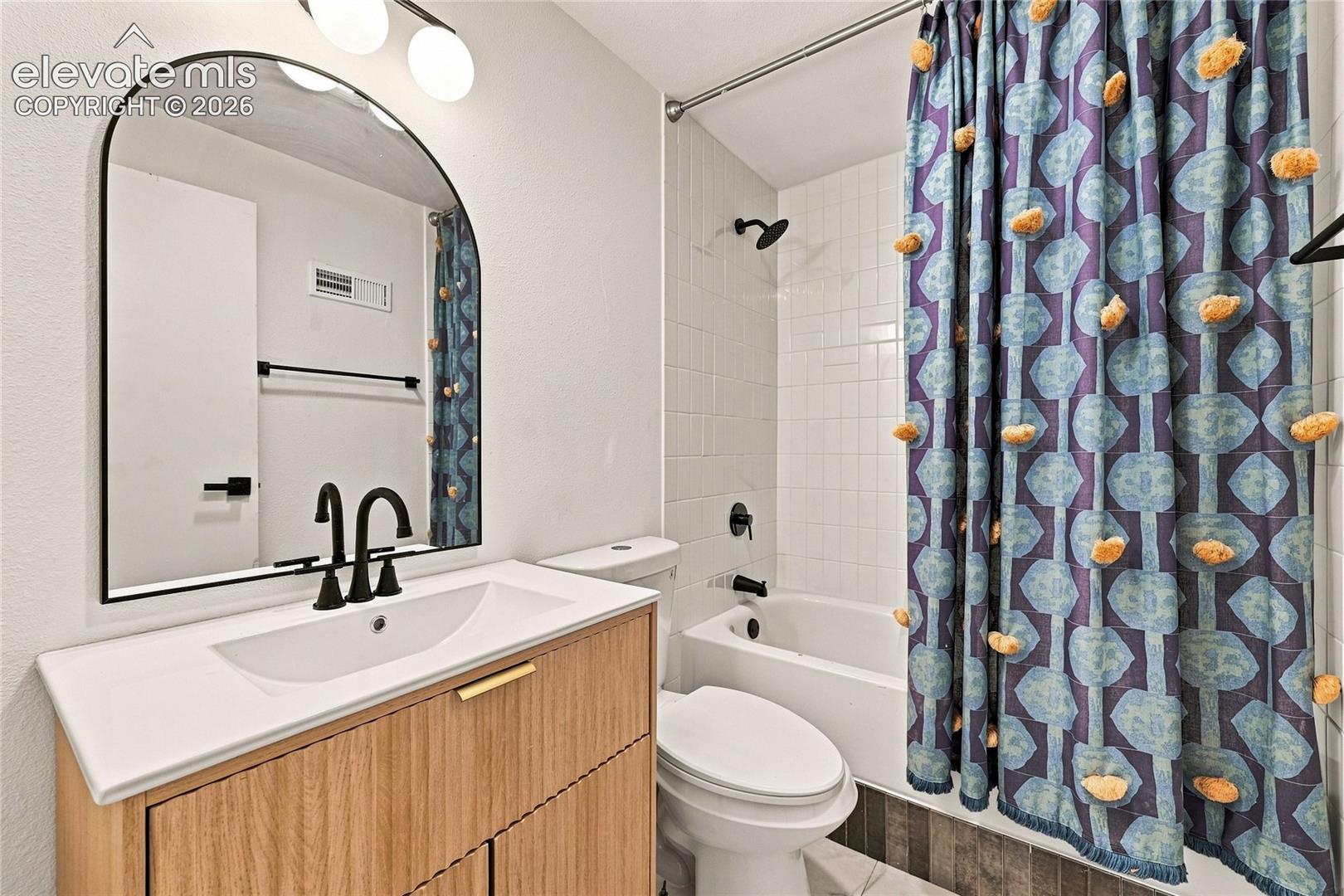 Bathroom featuring a wood-finish vanity with an integrated sink, black hardware, an arched mirror, and a tiled shower with a tub