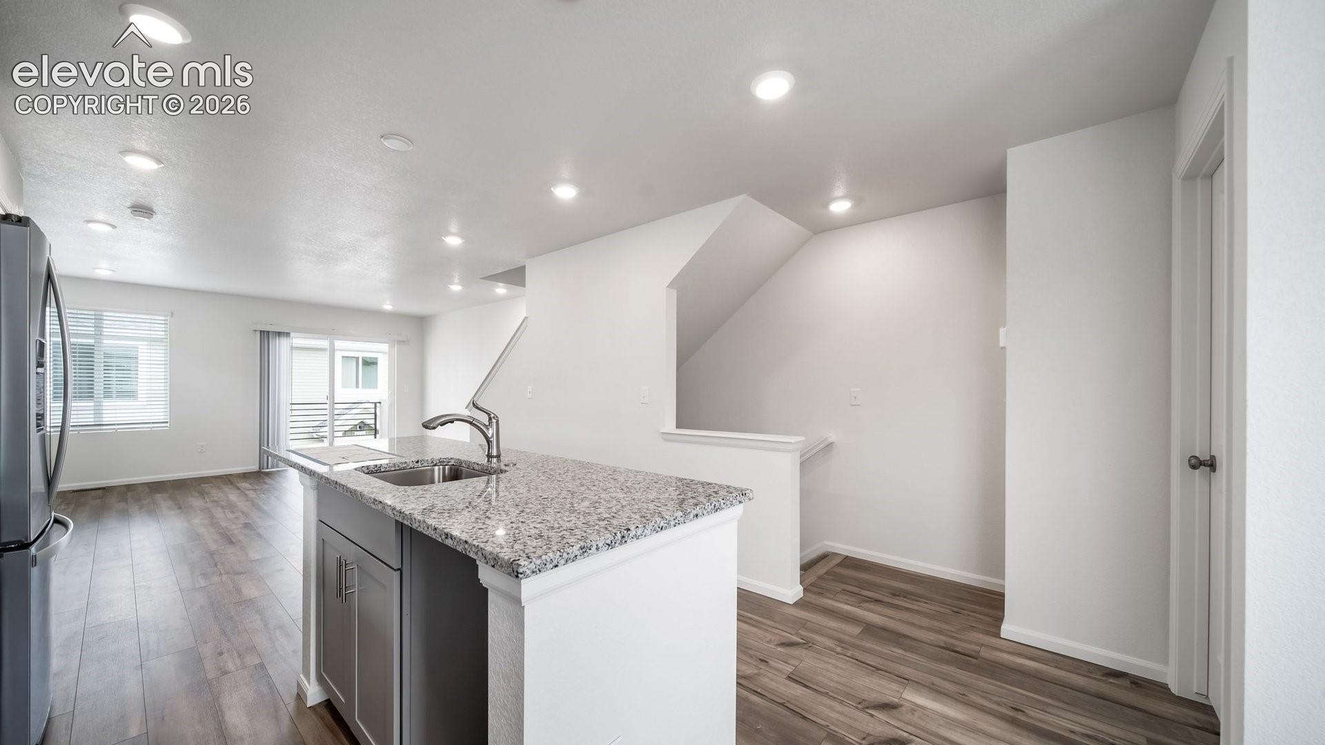 Kitchen island with a stainless steel sink and granite-finish countertop, stainless steel refrigerator, recessed lighting, wood-finish flooring, and white walls
*photos of a model home*