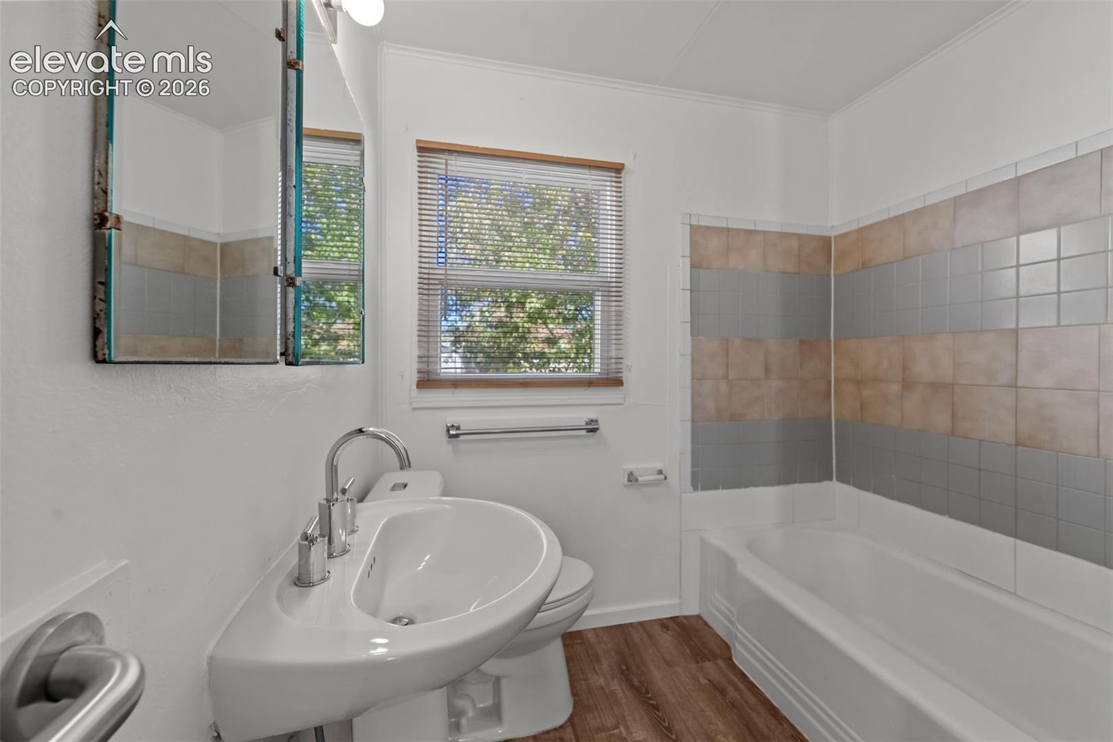 Wall-mounted sink with a curved basin and chrome faucet, wood-finish flooring, and a bathtub with decorative two-tone horizontal wall tile