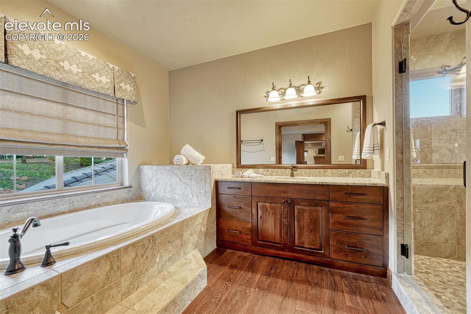Built-in soaking tub, wood-finish flooring, dark wood vanity with ample storage, tiled shower, and a large vanity mirror