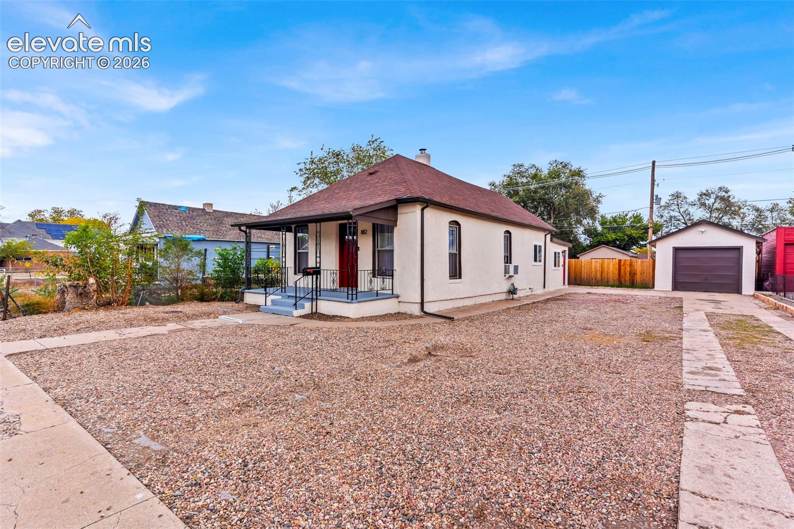 Stucco exterior featuring a covered front porch, arched window detailing, a dedicated detached garage, and an expansive gravel driveway