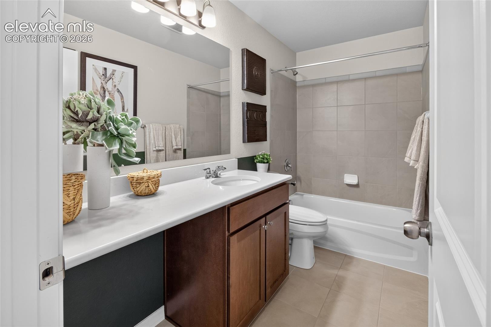 Bathroom featuring a wood-finish vanity with a white countertop, an integrated sink, a large wall-mounted mirror with multi-bulb lighting, and a tub-shower combination with light-toned wall tiling
