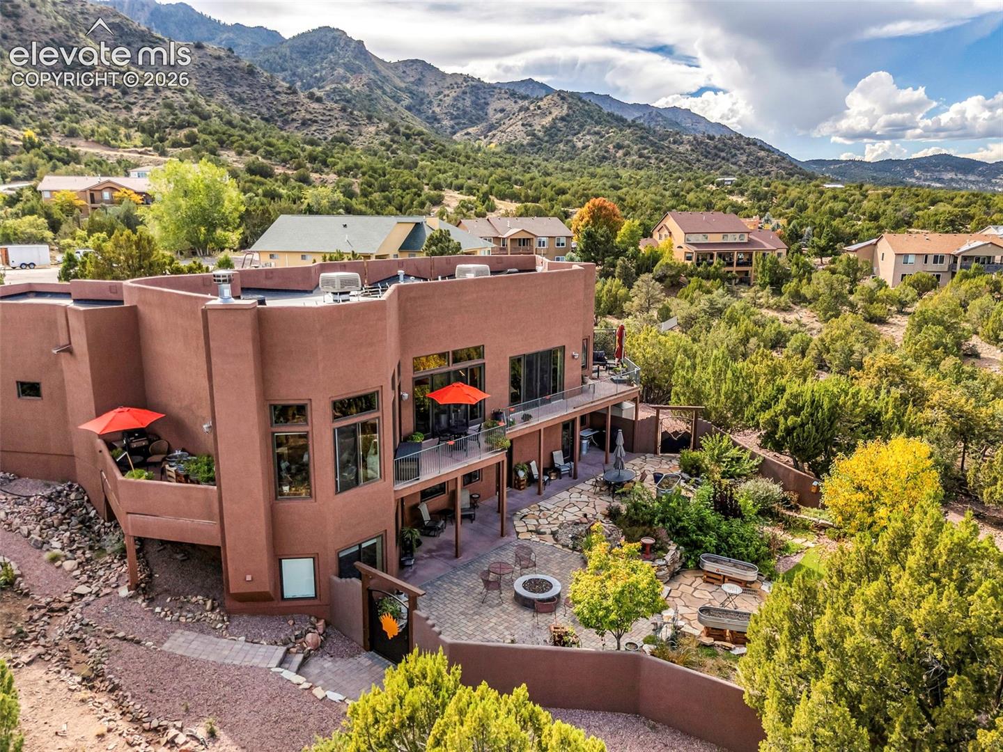 Multi-level stucco exterior featuring tiered balconies with metal railings, a stone-paved patio, an integrated fire pit, and a backdrop of mountain terrain