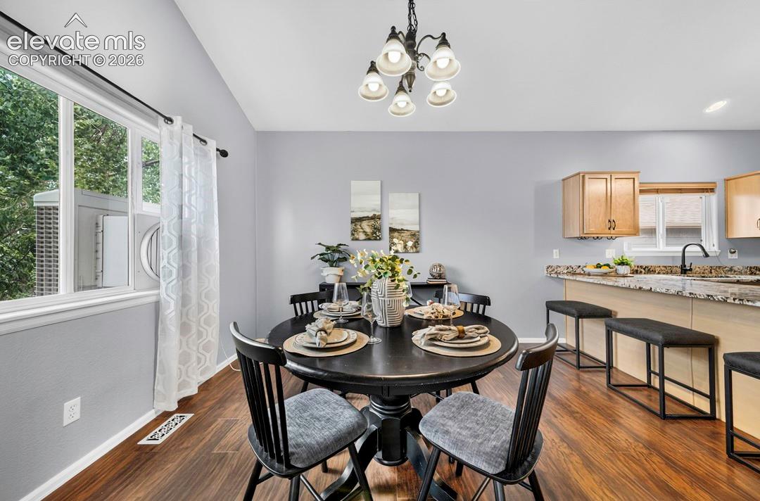 Open-concept dining area featuring vaulted ceilings, wood-finish flooring, a multi-bulb chandelier, and a kitchen island with stone-surface countertops