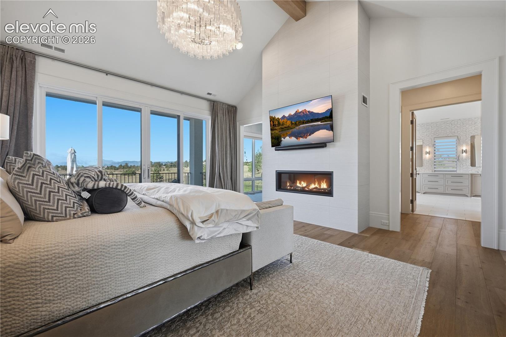Vaulted ceiling with exposed beam, linear fireplace, floor-to-ceiling large-format tile surround, wood-finish flooring, and multi-panel glass door system