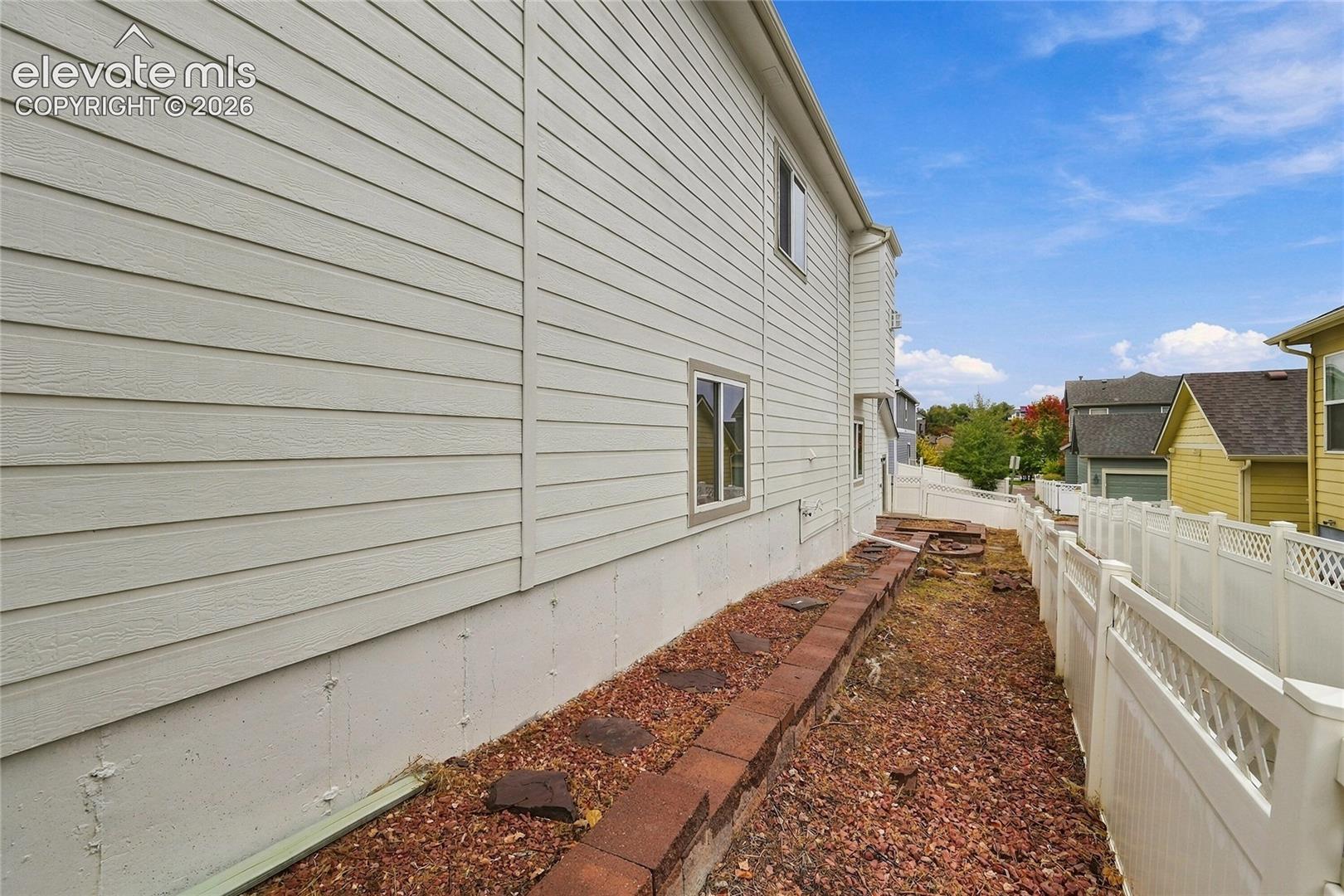 Exterior side elevation featuring horizontal siding, a concrete foundation, and a gravel ground cover with a segmental block retaining wall and vinyl privacy fencing