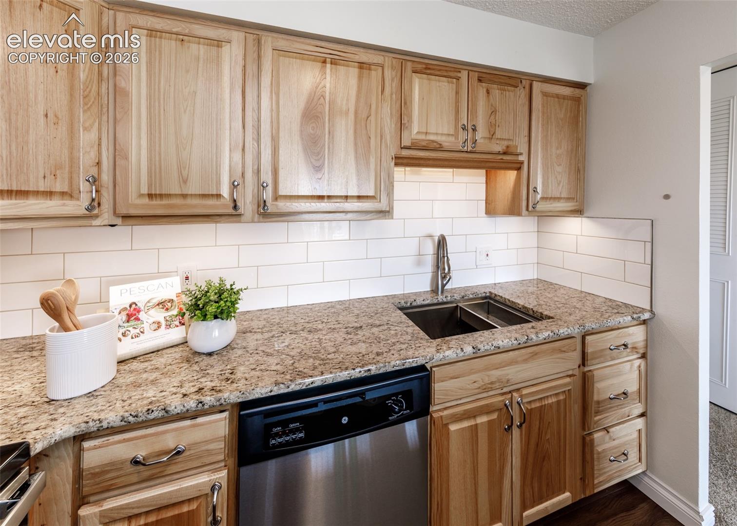 Natural wood-finish cabinetry, stone-surface countertops, white subway tile backsplash, stainless steel under-mount double basin sink, and built-in stainless steel dishwasher