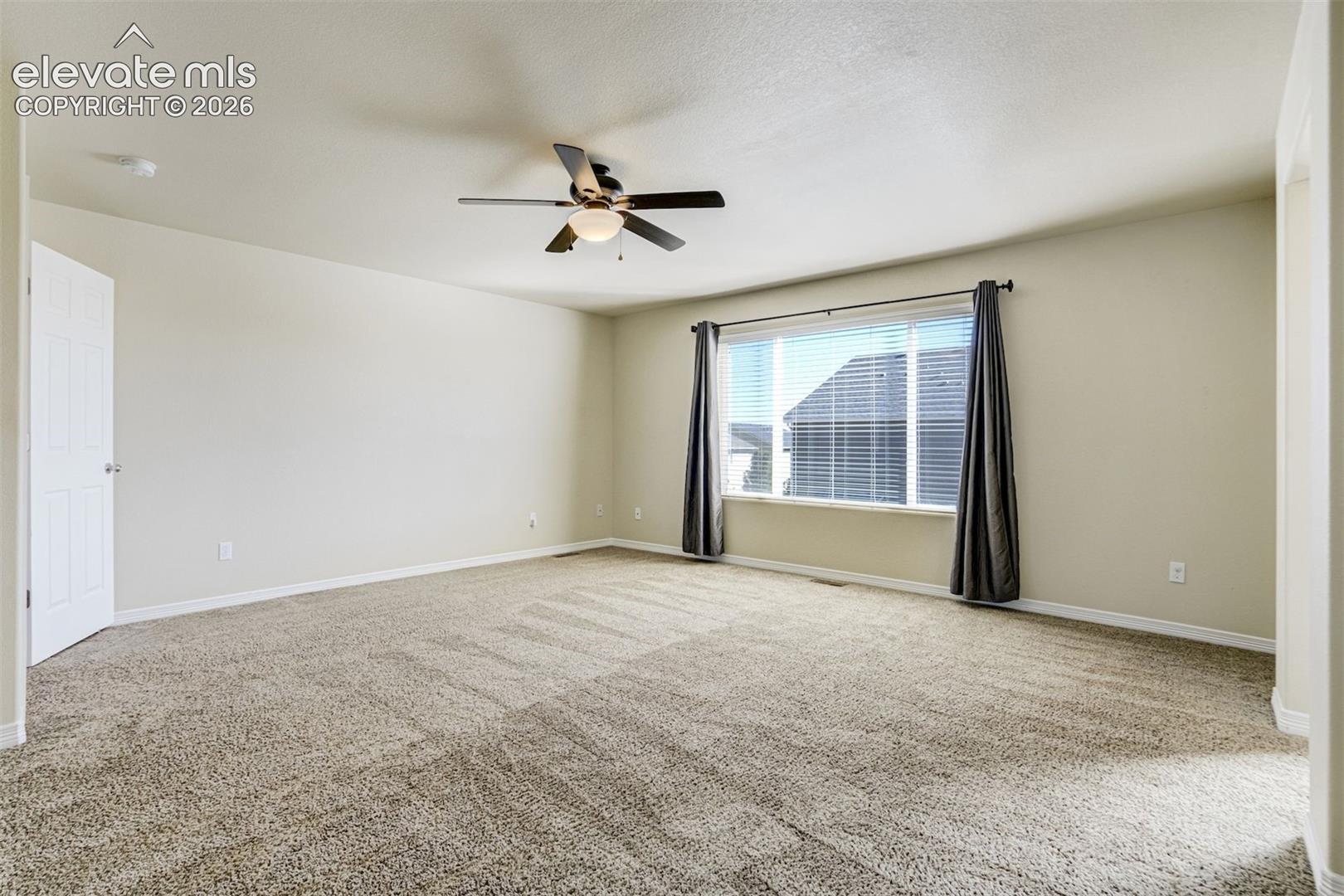 Spacious primary living area featuring neutral carpeting, ceiling fan with integrated lighting, large windows with blackout curtains, and abundant natural light from dual-pane glass