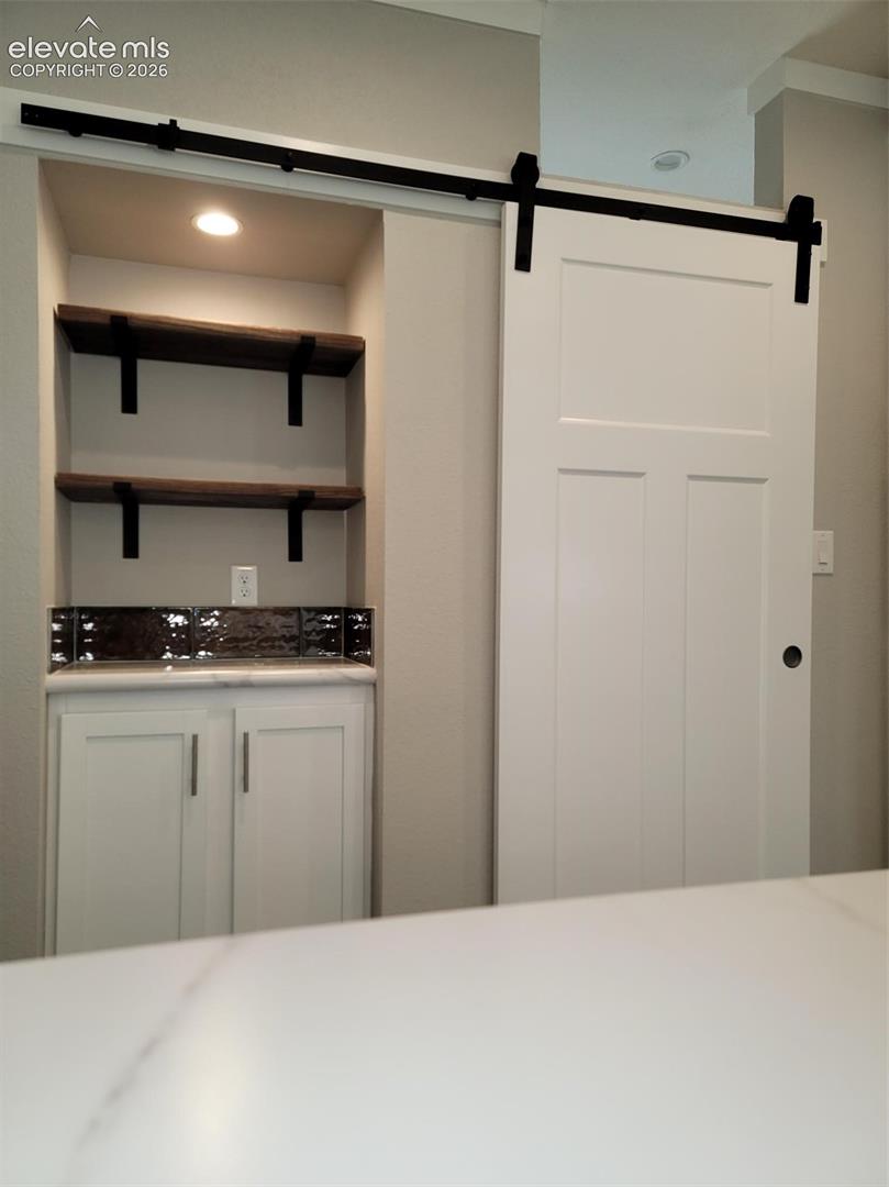 Built-in cabinetry with  countertop, dark backsplash, recessed lighting, and wooden shelving, paired with a white sliding barn door on matte black hardware
