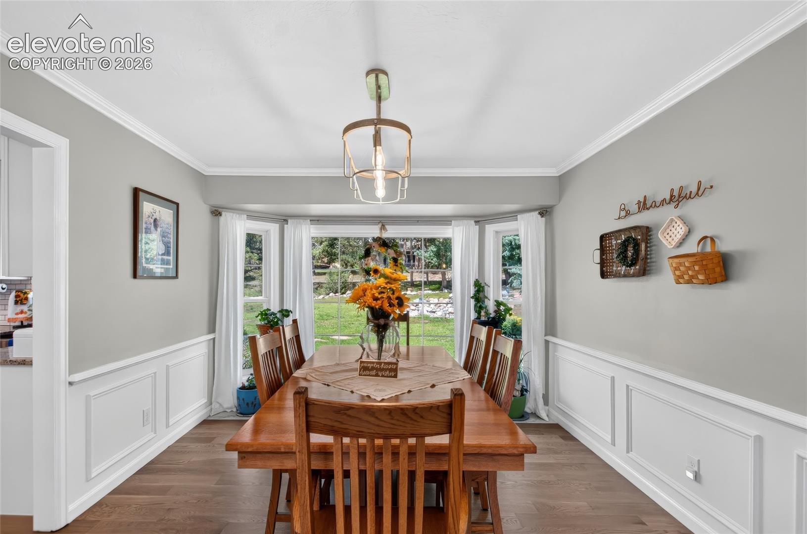 Bay window overlooking the yard, wood-finish flooring, wainscoting, crown molding, and a suspended light fixture