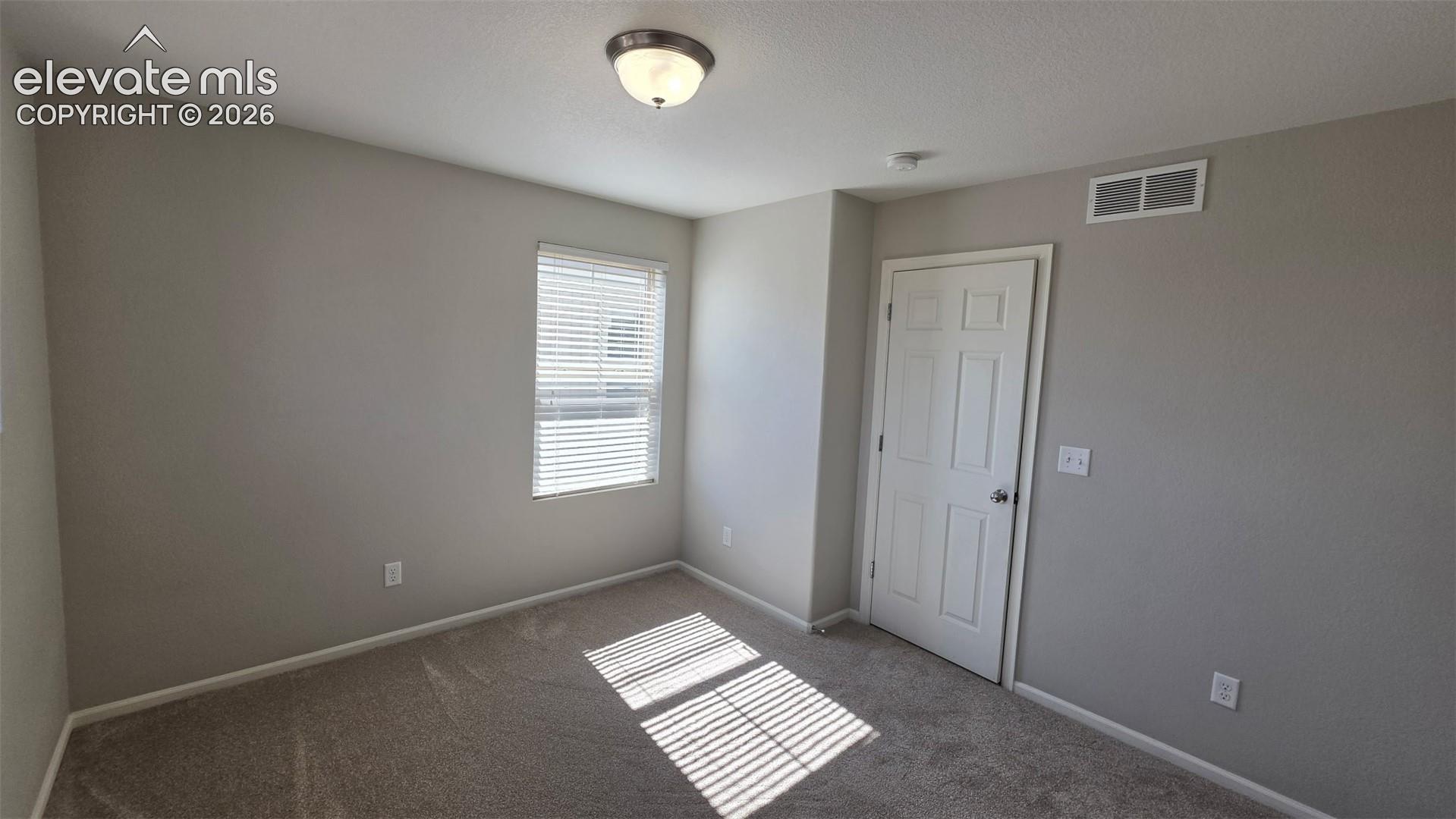 Neutral-toned interior space featuring wall-to-wall carpeting, a single-window with horizontal blinds, a six-panel door, and a flush-mount ceiling light fixture